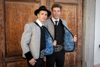 Madratzengradl Tuxer Janker Zwei Männer in Tuxer Tracht vor einer geschnitzten Holztür, die Lodenjanker offen, das hellblaue Futter sichtbar.