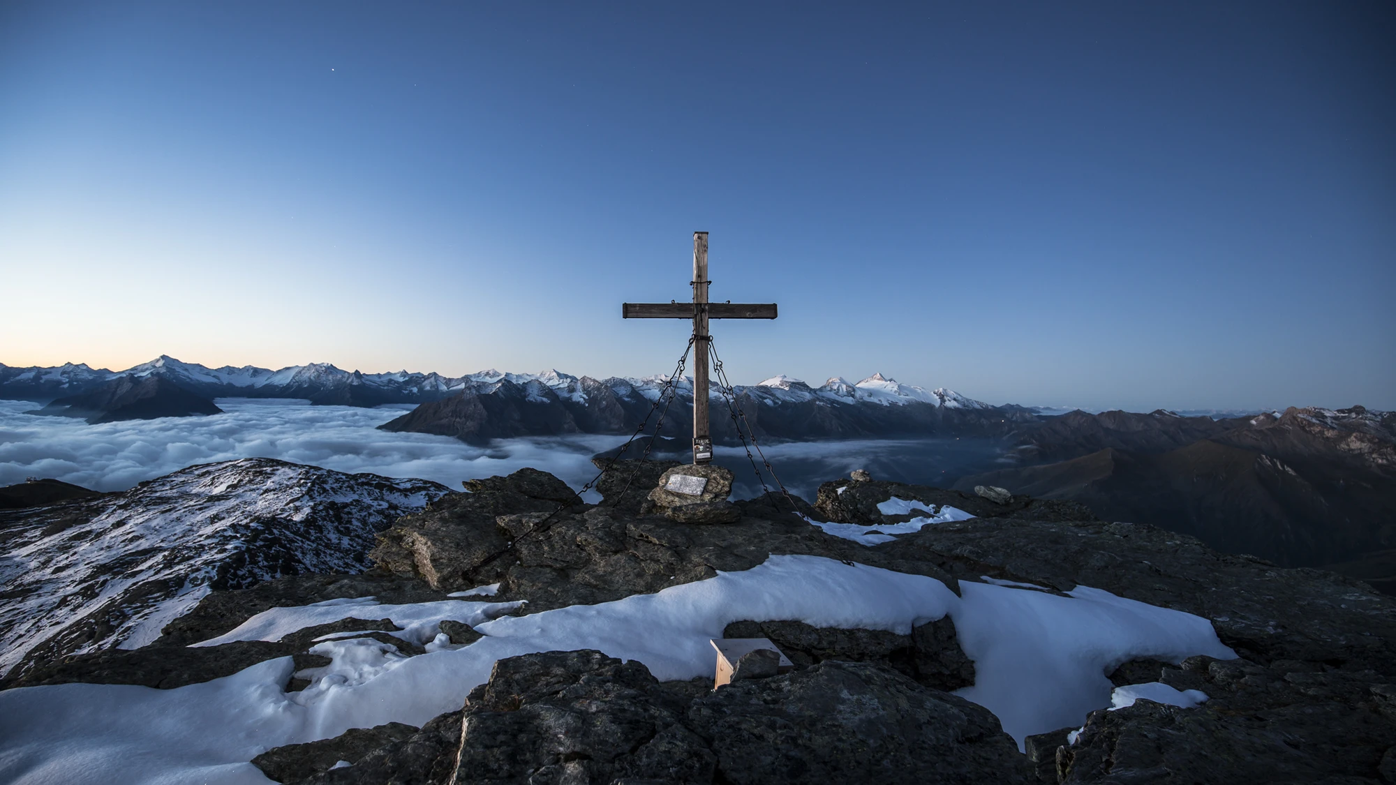 Holzkreuz am Gipfel des Rastkogels in Tux, mit Eisenketten verankert, darunter Wolkenmeer, im Hintergrund die Zillertaler Alpen.