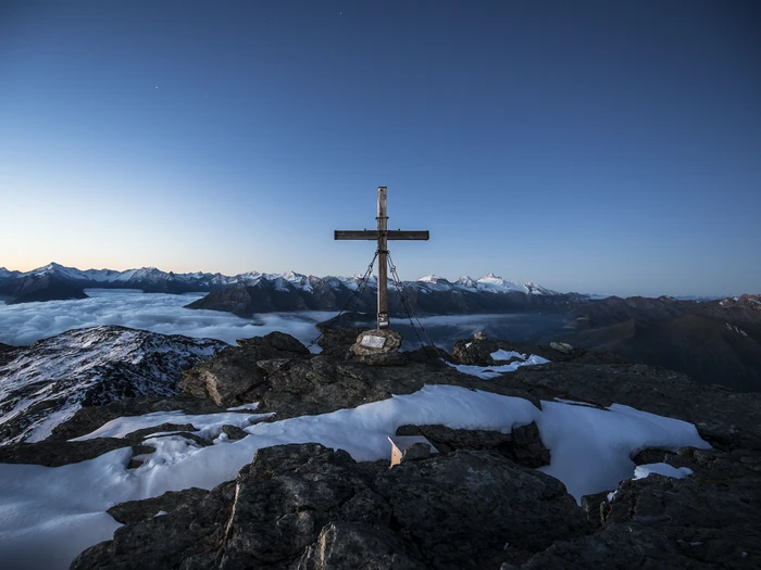 Gipfelkreuz Rastkogel Holzkreuz am Gipfel des Rastkogels in Tux, mit Eisenketten verankert, darunter Wolkenmeer, im Hintergrund die Zillertaler Alpen.