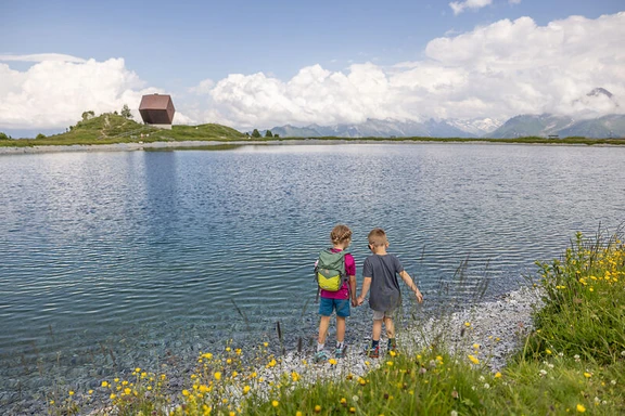 Penkensee Zwei Kinder stehen am Ufer des Penkensees im Zillertal, dahinter eine Hütte, Blumenwiese im Vordergrund.