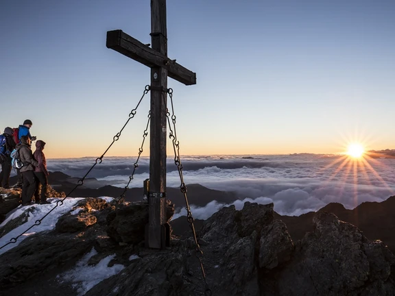 Rastkogel bei Sonnenaufgang Gipfelkreuz am Rastkogel in Tux bei Sonnenaufgang, daneben Wanderer im Schnee, darunter ein Wolkenmeer.