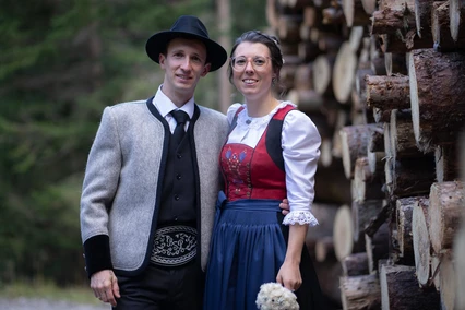 Tuxer Tracht Paar in Tuxer Tracht vor gestapeltem Rundholz: grauer Lodenjanker mit Federkielgürtel, rotes besticktes Mieder, blaue Schürze.