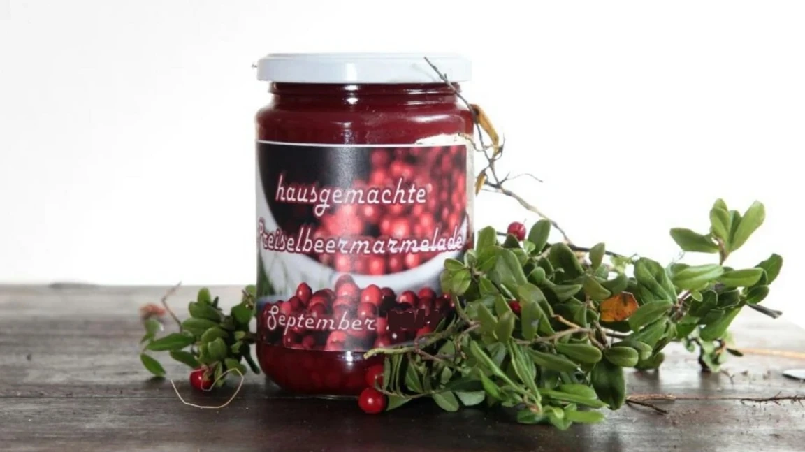 Preiselbeermarmelade aus dem Hotel Eden Glas hausgemachte Preiselbeermarmelade mit handbeschriftetem Etikett, daneben Preiselbeerzweige auf einem Holztisch.